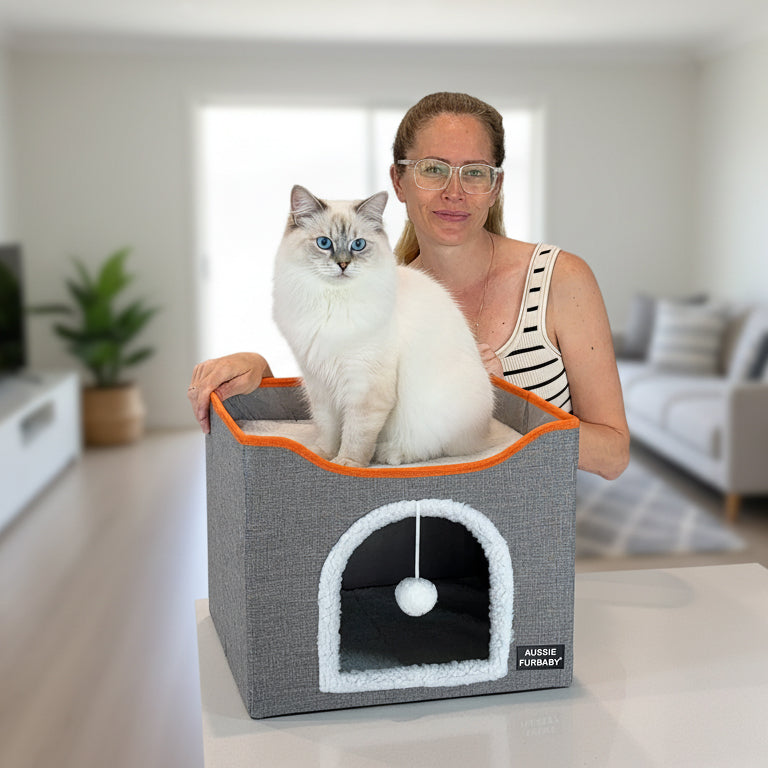 Woman holding a cat in a gray pet bed with orange trim in a living room setting.